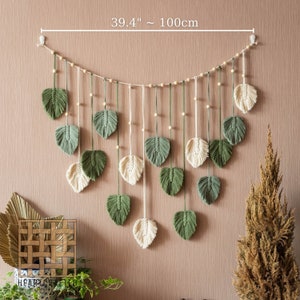 May include: A macrame wall hanging with green and cream leaf shapes, measuring 39.4 inches or 100 centimeters in length. The hanging is made of cotton cord and features wooden beads.
