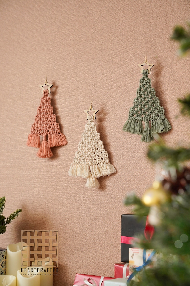 K&ouml;nnte beinhalten: Drei Makramee-Weihnachtsbaumschmuck mit Holzstern-Toppern. Die B&auml;ume sind in den Farben Gr&uuml;n, Beige und Koralle gehalten. Der Schmuck h&auml;ngt an einer beigefarbenen Wand. Der Text "HEARTCRAFTED" ist unten im Bild zu sehen.