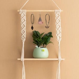 May include: A wooden shelf with two tiers, decorated with macrame hanging planters. The shelf has three hooks for hanging jewellery. A potted plant sits on the top shelf.