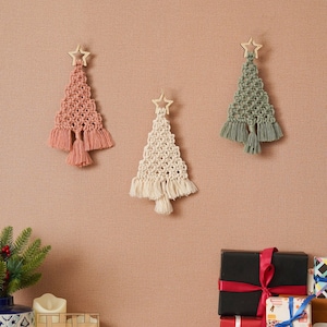 K&ouml;nnte beinhalten: Drei Makramee-Weihnachtsbaumschmuck mit Holzstern-Toppern. Die B&auml;ume sind rosa, wei&szlig; und gr&uuml;n. Der Schmuck h&auml;ngt an einer braunen Wand. Im Vordergrund befinden sich verpackte Geschenke.