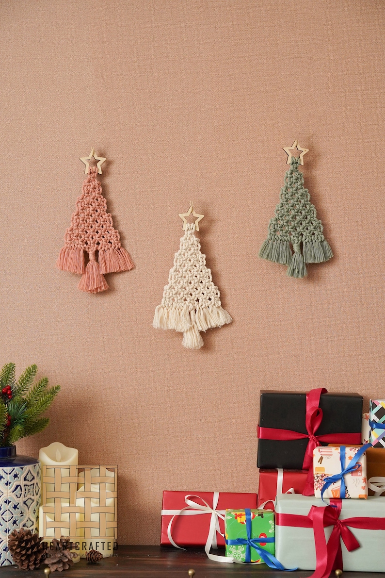 K&ouml;nnte beinhalten: Drei Makramee-Weihnachtsbaumschmuck mit Holzstern-Toppern. Die B&auml;ume sind rosa, wei&szlig; und gr&uuml;n. Der Schmuck h&auml;ngt an einer braunen Wand. Im Vordergrund befinden sich verpackte Geschenke.