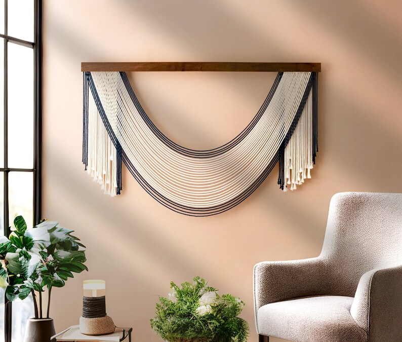 May include: A white and navy blue macrame wall hanging with a curved shape. The hanging is made of cotton cord and is attached to a wooden dowel.