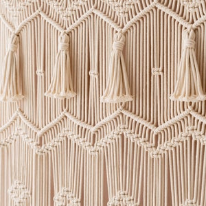 May include: A white macrame wall hanging with a geometric pattern and tassels. The hanging is made of cotton cord and has a natural, bohemian style.