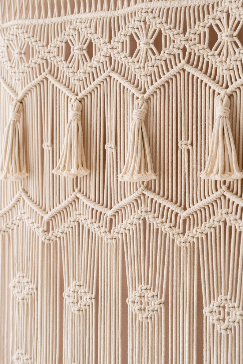 May include: A white macrame wall hanging with a geometric pattern and tassels. The hanging is made of cotton cord and has a natural, bohemian style.