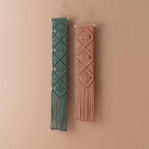 May include: Two macrame wall hangings, one in teal and one in blush pink, with a diamond pattern and fringe.