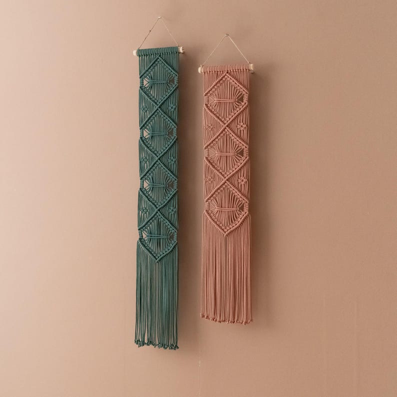 May include: Two macrame wall hangings, one in teal and one in blush pink, with a diamond pattern and fringe.