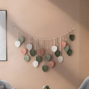 May include: A wall hanging made of yarn in the shape of leaves. The leaves are in shades of green, white, and pink. The leaves are strung together on a white string.