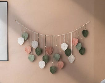 May include: A wall hanging made of yarn in the shape of leaves. The leaves are in shades of green, white, and pink. The leaves are strung together on a white string.