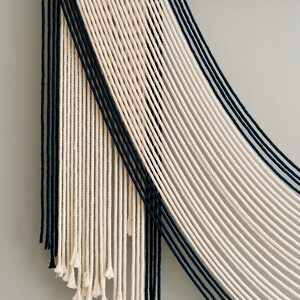 May include: A wall hanging made of white and black string, with a geometric pattern. The strings are woven together to create a modern and abstract design.