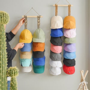 May include: A display of baseball caps in various colors, including yellow, blue, and green, hanging on a wall. The caps are organized on wooden rods suspended by woven rope. A person is seen adjusting one of the displays.