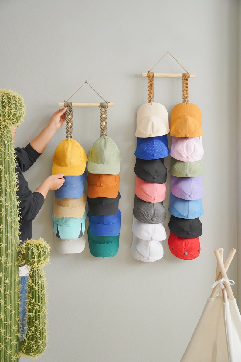 May include: A display of baseball caps in various colors, including yellow, blue, and green, hanging on a wall. The caps are organized on wooden rods suspended by woven rope. A person is seen adjusting one of the displays.