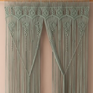 May include: A macrame wall hanging in a soft sage green color. The design features intricate geometric patterns and long, flowing fringe. The piece is suspended from a wooden dowel, adding a natural touch to the handcrafted textile art.