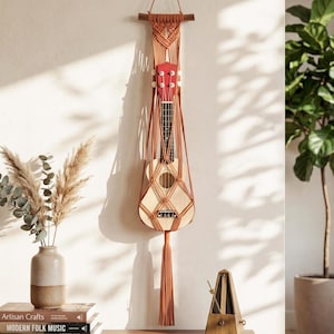 May include: A ukulele with a red headstock is suspended in a macrame plant hanger. The hanger is a warm orange color and is attached to a wooden dowel. The ukulele has a natural wood finish and is hanging on a white wall.