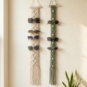 May include: Two macrame sunglass holders in beige and olive green hang on a white wall. Each holder has multiple pairs of sunglasses displayed. The beige holder has a diamond pattern, while the green holder has wooden beads.