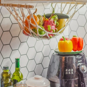 May include: A white hammock hanging from a shelf, filled with fresh fruit, including a dragon fruit, a yellow mango, a green cucumber, and a few green apples.  A black air fryer sits on a countertop below the hammock, with two bell peppers on a plate on top of the air fryer.  Two bottles of olive oil are on the counter, one with a green label that says "Extra Virgin Olive Oil" and the other with a white label that says "Artcraft".  A white rice cooker is also on the counter.