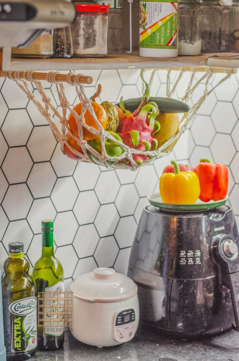 May include: A white hammock hanging from a shelf, filled with fresh fruit, including a dragon fruit, a yellow mango, a green cucumber, and a few green apples.  A black air fryer sits on a countertop below the hammock, with two bell peppers on a plate on top of the air fryer.  Two bottles of olive oil are on the counter, one with a green label that says "Extra Virgin Olive Oil" and the other with a white label that says "Artcraft".  A white rice cooker is also on the counter.