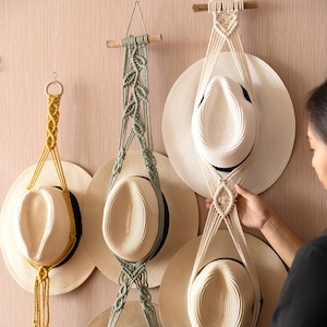 Puede incluir: Tres colgadores de macramé para sombreros en amarillo mostaza, verde salvia y blanco crema, con diseños de cordón anudado suspendidos de varillas o anillos de madera, sosteniendo sombreros de paja de ala ancha con cintas negras en vertical.