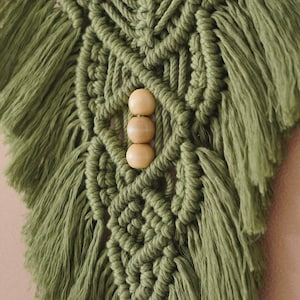 May include: A green macrame wall hanging with wooden beads. The wall hanging has a geometric pattern and is made with thick yarn.