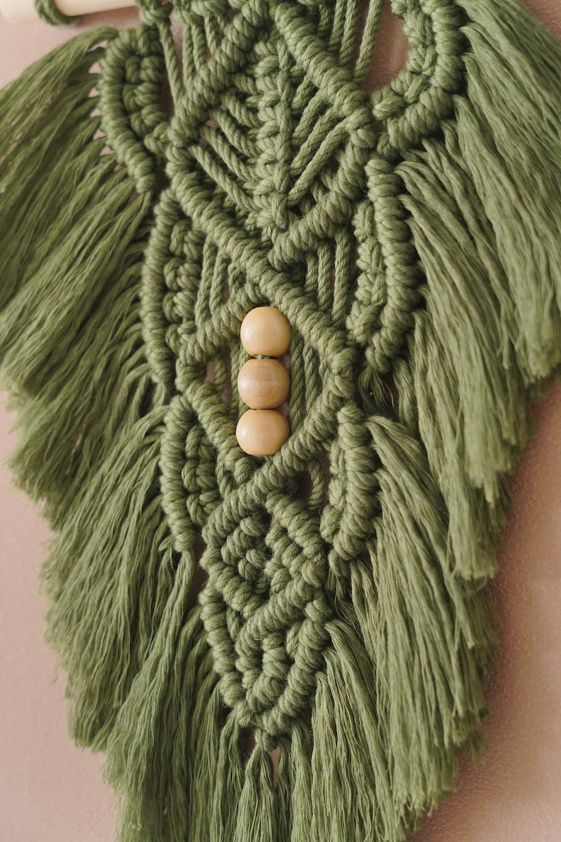 May include: A green macrame wall hanging with wooden beads. The wall hanging has a geometric pattern and is made with thick yarn.