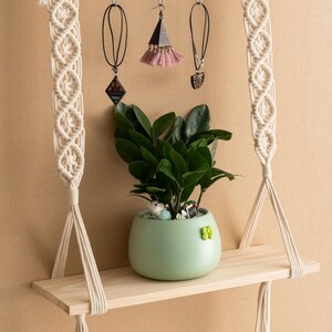 May include: A wooden shelf with macrame hanging cords, a small potted plant, and three hanging jewellery pieces. The shelf is suspended from the ceiling by the macrame cords.