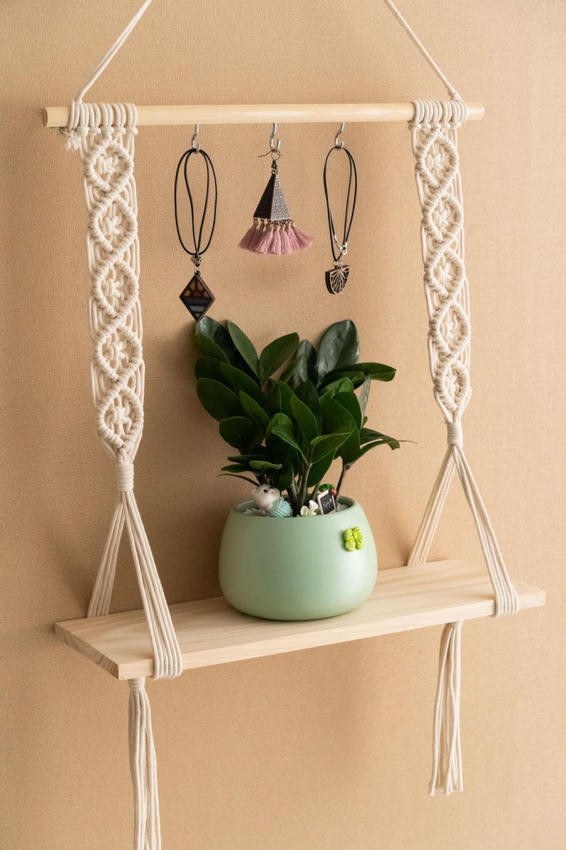 May include: A wooden shelf with macrame hanging cords, a small potted plant, and three hanging jewellery pieces. The shelf is suspended from the ceiling by the macrame cords.