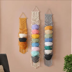 May include: Three macrame wall organizers, each with multiple baseball caps in various colors. The organizers are in mustard yellow, off-white, and gray. The caps include shades of pink, yellow, blue, and green, creating a colorful display.