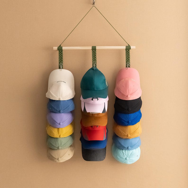 May include: A wooden hat rack with three rows of baseball caps in various colors, including white, blue, pink, and green. The rack is suspended from a wall by a green cord. The caps are neatly stacked, showcasing a practical storage solution for headwear.