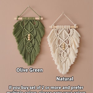 May include: Two macrame wall hangings, one in olive green and one in natural white, with wooden dowels and beads. The hangings are in the shape of a leaf.