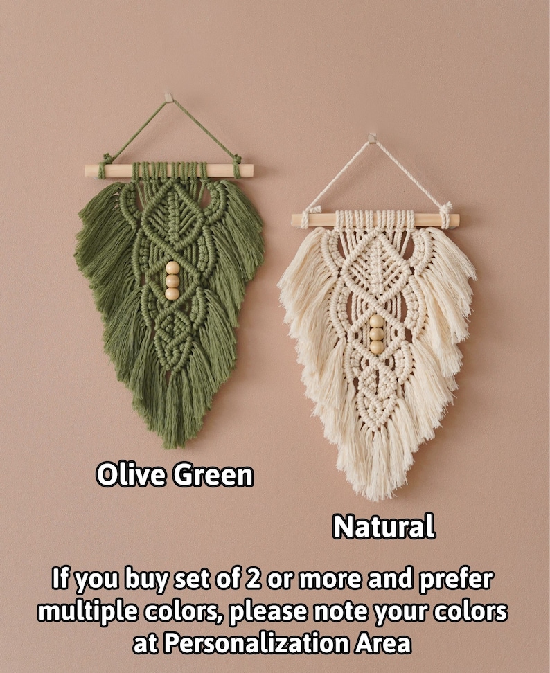 May include: Two macrame wall hangings, one in olive green and one in natural white, with wooden dowels and beads. The hangings are in the shape of a leaf.