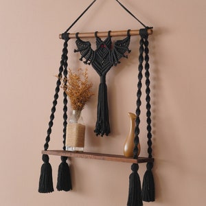 May include: Black macrame wall shelf with a bat design and black tassels. The shelf is made of wood and has a natural finish.