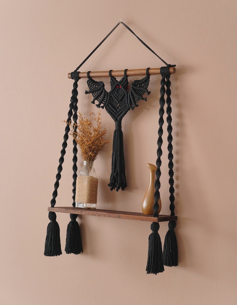 May include: Black macrame wall shelf with a bat design and black tassels. The shelf is made of wood and has a natural finish.