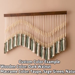 May include: A wall hanging with a wooden frame and a curved design made of white cord, wooden beads, and tassels in taupe, sage green, and natural colors. Custom Color Example + Wooden Color: Dark Walnut + Macrame Color: Taupe, Sage Green, Natural