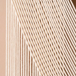 May include: A white macrame wall hanging with a geometric pattern. The macrame is made with thick cotton cord and is attached to a wooden frame.
