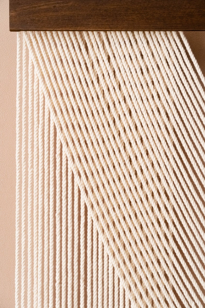 May include: A white macrame wall hanging with a geometric pattern. The macrame is made with thick cotton cord and is attached to a wooden frame.