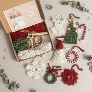 Può includere: Un kit artigianale con il testo "Basic Knots" e vari materiali per macramè, tra cui cordoncini rossi, verdi e bianchi, perline di legno e un ornamento a forma di stella. Sono esposti anche diversi ornamenti natalizi in macramè fatti a mano.