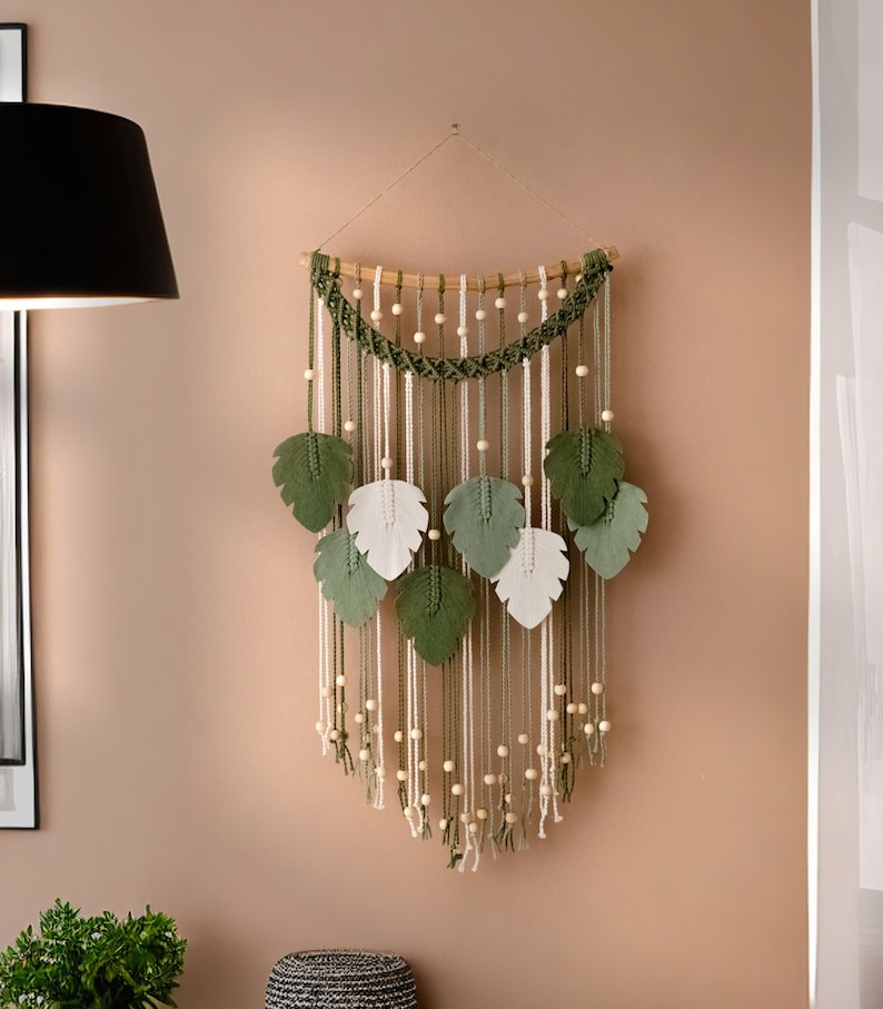 Op de afbeelding: Een macram&eacute; wandhanger met groene en witte viltbladeren en houten kralen. De hanger is gemaakt van natuurlijke vezels en heeft een bohemian stijl.