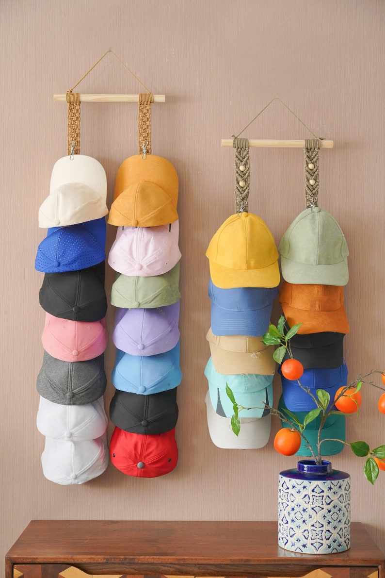 May include: Two macrame hat racks with a variety of baseball caps in different colors. The hats are hanging in vertical rows. A decorative vase with a branch of oranges sits on a wooden surface.