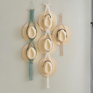 May include: Three macrame wall hangers made of cotton cord, designed to hold hats. One hanger is teal and holds two straw hats, one is off-white and holds three straw hats, and one is beige and holds a single straw hat with a black ribbon band.
