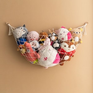 Op de afbeelding: Een witte macramé speelgoedhangmat gevuld met diverse knuffels. De knuffels omvatten een grijze uil, een roze zeester, een witte kat met roze accenten en een bruine beer. De hangmat hangt tegen een beige muur.