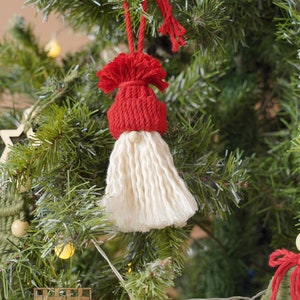 May include: A red and white yarn gnome ornament hanging on a Christmas tree. The gnome has a red hat with a white pom-pom and a long white beard. The gnome is hanging from a string.