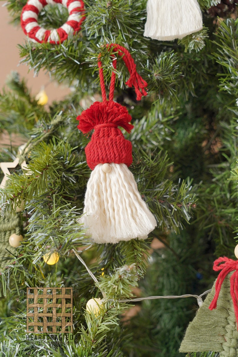 May include: A red and white yarn gnome ornament hanging on a Christmas tree. The gnome has a red hat with a white pom-pom and a long white beard. The gnome is hanging from a string.