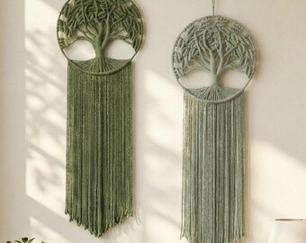 May include: Two macrame wall hangings featuring a tree of life design. One is olive green, the other is a muted sage green. Each has a circular frame with a detailed tree and long, fringed tassels. The wall hangings are suspended against a neutral wall.