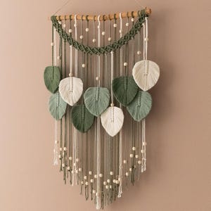Può includere: Un arazzo da parete in macramè con uno schema di colori verde e bianco. L'arazzo presenta una serie di foglie intrecciate e perline.