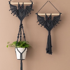 May include: Two dark grey macrame plant hangers, one holding a white pot with a green plant. The macrame design resembles a bat with outstretched wings, suspended from a wooden dowel. The other hanger is a similar design, without a pot.