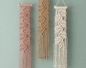 May include: Three vertical macrame wall hangings suspended from wooden dowels with string loops. Each piece features a repeating knotted leaf pattern in soft pink, tan, and cream colours, ending in long, loose fringe with small knotted accents.