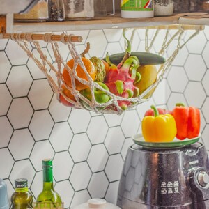 May include: A white string hammock hanging from a wooden shelf, filled with fresh fruit, including a dragon fruit, a green apple, a yellow mango, and an orange.  The hammock is hanging over a white hexagonal tiled wall.  A black air fryer and a white rice cooker are on the counter below the hammock.