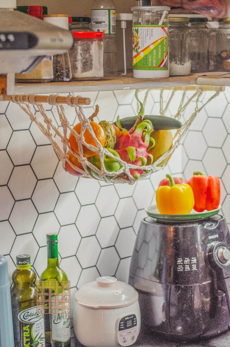 May include: A white string hammock hanging from a wooden shelf, filled with fresh fruit, including a dragon fruit, a green apple, a yellow mango, and an orange.  The hammock is hanging over a white hexagonal tiled wall.  A black air fryer and a white rice cooker are on the counter below the hammock.