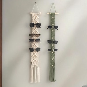 Puede incluir: Dos organizadores de gafas de sol de macramé colgando de una pared. Uno es blanquecino, el otro verde oliva. Cada uno sostiene varios pares de gafas de sol. El organizador blanquecino tiene un patrón de diamantes, mientras que el verde tiene un diseño vertical con cuentas de madera.
