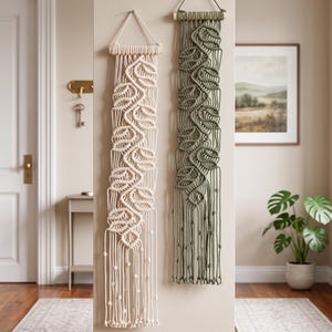 Puede incluir: Dos tapices de macramé verticales colgados de varillas de madera. Una pieza es de color crema y la otra es verde oliva. Ambas presentan intrincados patrones de nudos en forma de hoja con flecos largos colgantes y detalles de nudos.