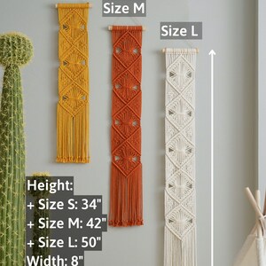 May include: Three macrame wall hangings in different sizes, with a yellow, orange, and white color scheme. The hangings are made with a knotting technique and have a fringe at the bottom. The sizes are labeled as "Size S", "Size M", and "Size L". The measurements are listed as: Height: Size S: 34", Size M: 42", Size L: 50", Width: 8".